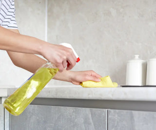 En persons händer rengör en köksbänk med en sprayflaska och en gul trasa.