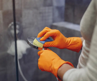 Händer i orange handskar som rengör ett duschhuvud med en svamp.